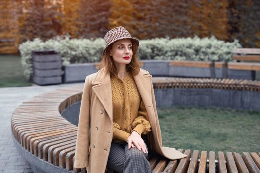 Charming young woman in beige coat and plaid hat sits on round bench waiting for her date or girlfriends. Autumn walk concept. Tinted photo.