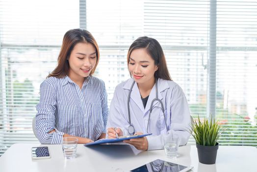 Female doctor talks to female patient in hospital office while writing on the patients health record on the table. Healthcare and medical service. 