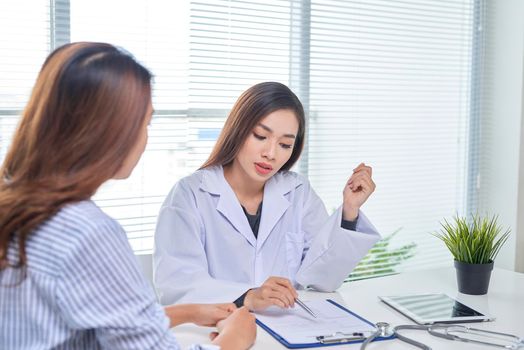 Female doctor talks to female patient in hospital office while writing on the patients health record on the table. Healthcare and medical service. 