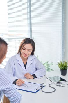 Female doctor talks to female patient in hospital office while writing on the patients health record on the table. Healthcare and medical service. 