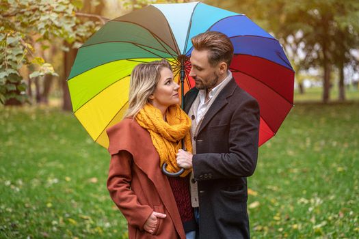 Beautiful in love couple standing in the park under a rainbow colored umbrella looking at each others eyes. A beautiful girl walks through the autumn park in rainy weather. 