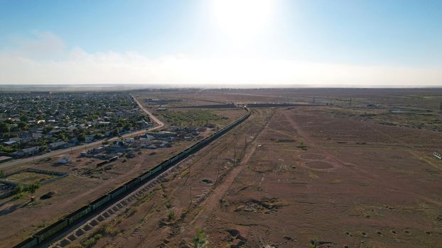 The train is traveling by rail. Railway station. Freight train and passenger train. There are large wagons and containers. Top view from the drone. Blue sky, small buildings and a road. Balkhash