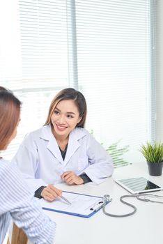 Female doctor talks to female patient in hospital office while writing on the patients health record on the table. Healthcare and medical service. 