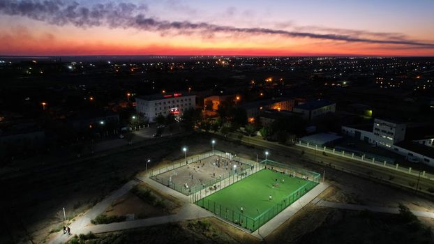 A group of people are playing football and basketball on the playground. The playground is located in the park. Lanterns are lit, people are resting and having fun. Sunset over the city. Kazakhstan