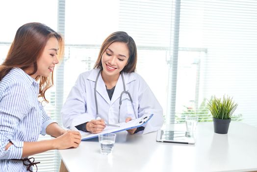 Female doctor talks to female patient in hospital office while writing on the patients health record on the table. Healthcare and medical service. 