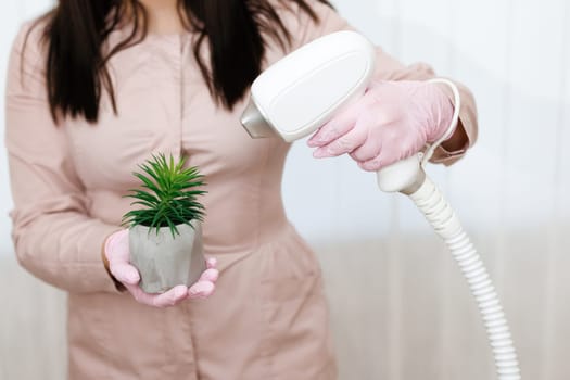 Woman's hand holding a vase in a gray pot. Laser device for hair removal is directed at it on the background of a person. Hair removal, fun, laser hair removal, fun.