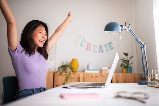 Excited and happy young teen asian female raising arms up celebrating success or achievement at home using laptop. Victory concept.