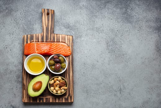 Food sources of healthy unsaturated fat and omega 3: fresh raw salmon fillet, avocado, olives, nuts on cutting board, rustic stone background top view. Healthy nutrition and keto diet, space for text