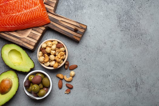 Food sources of healthy unsaturated fat and omega 3: fresh raw salmon fillet, avocado, olives, nuts on cutting board, rustic stone background top view. Healthy nutrition and keto diet, space for text