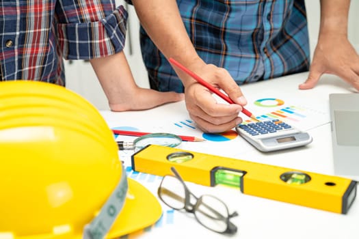 Asian engineer and partner team learn and talk with house plan project blueprint, graph and yellow helmet and engineering construction tools on table work.