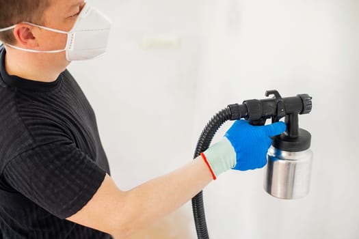 Worker in protective mask painting wall with spray gun. Painting the wall of apartment. Repair concept