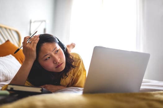 Stressed, worried Asian teen girl lying on bed attending online classes listening teacher lesson with headphones using laptop. Doing homework. E-learning concept.