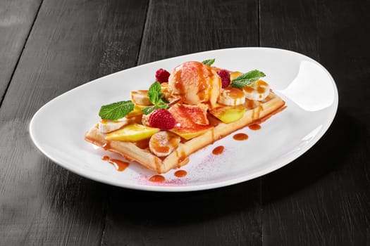 Delicious Belgian waffle topped with sweet banana and mango slices, ripe raspberries and scoop of vanilla ice cream, seasoned with caramel sauce and garnished with fresh mint leaves. Popular dessert