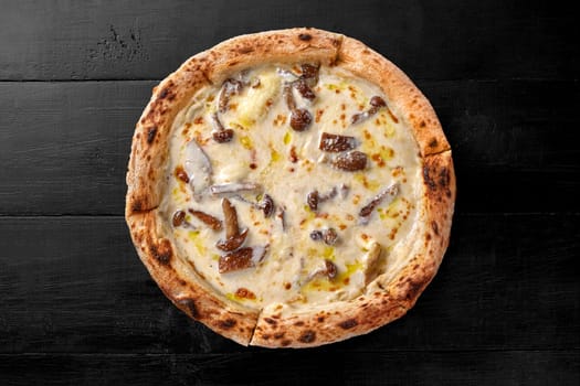 Top view of sliced delicious pizza with delicate creamy cheese sauce and fried forest mushrooms on black wooden surface. Popular Italian style food
