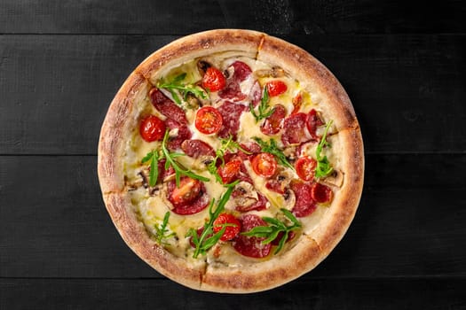 Delicious freshly baked Italian Boscaiola pizza with cream sauce, mozzarella, slices of cabanossi and salami, mushrooms, parmesan garnished with fresh arugula leaves on black wooden surface