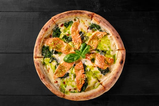 Appetizing pizza with browned edge filled with cheese sauce, slices of salmon and spinach dressed with fragrant pesto and fresh basil leaves on black wooden background. Italian style snack