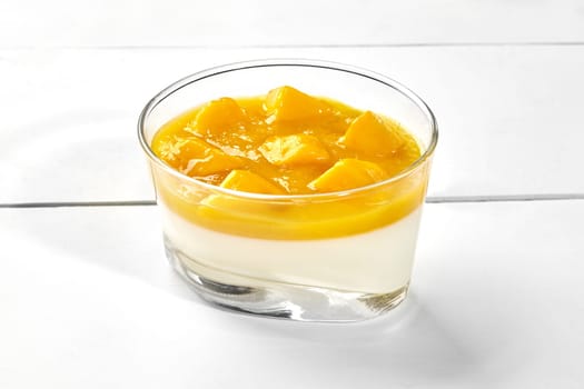 Delicious Italian creamy vanilla panna cotta with layer of mango puree and pieces of fresh ripe fruits served in glass on white wooden background. Popular desserts concept