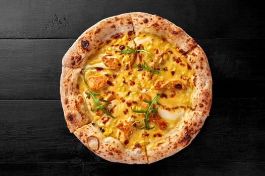 Appetizing freshly baked pizza with melted cheese mixture, sous vide chicken filet and corn sprinkled with fragrant arugula on black wooden background, top view
