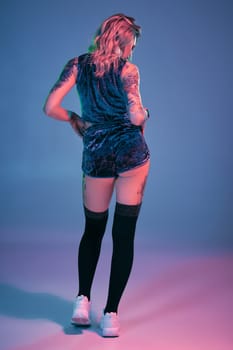 Photo of beautiful young blonde fitness girl in velour blue sport wear and black stockings seductively posing on camera in studio. Pink and blue background. Fitness, sport, twerk, dancing