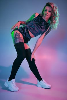 Photo of beautiful young blonde fitness girl in velour blue sport wear and black stockings seductively posing on camera in studio. Pink and blue background. Fitness, sport, twerk, dancing