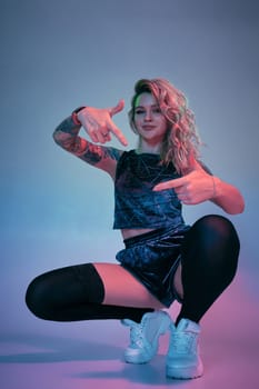 Beautiful young blonde tattoed girl in blue velour booty shorts and t-shirt, black stockings, squats in front of the camera in a confident pose. Pink and blue background