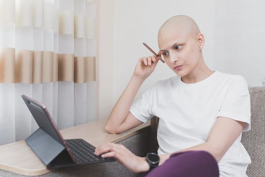 Hairless young woman with cancer wearing a white t-shirt working at laptop in room.