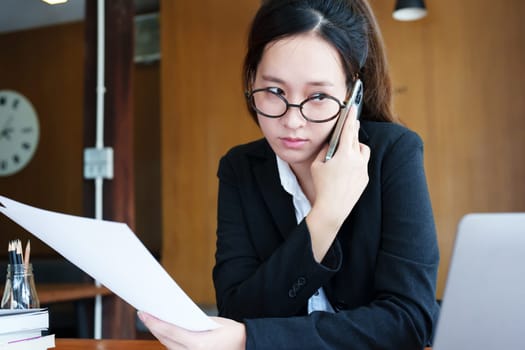 Portrait of a young Asian woman showing a serious face as she uses her phone, financial documents and computer laptop on her desk in the early morning hours.