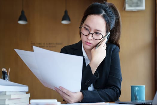 Portrait of a young Asian woman showing a serious face as she uses her phone, financial documents and computer laptop on her desk in the early morning hours.