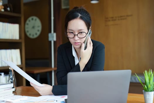 Portrait of a young Asian woman showing a serious face as she uses her phone, financial documents and computer laptop on her desk in the early morning hours.