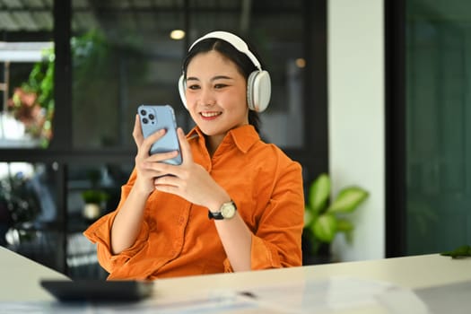 Cheerful stylish young woman wearing wireless headphone and using smartphone at her office desk.