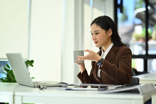 Beautiful female company worker or manager holding coffee cup and using laptop at office desk.