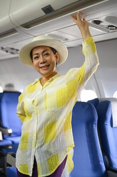 Portrait of 50s woman tourist standing at airplane aisle and smiling to camera. Retirement, travel and summer vacation concept.