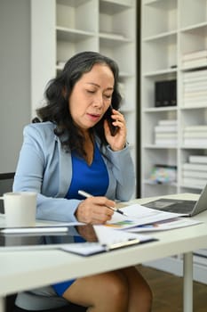 Beautiful mature businesswoman having pleased phone conversation and taking notes.