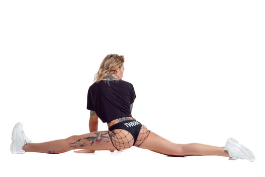 Nice blond twerk female with tattoed body and long curly hair is posing doing a twine isolated on white background with copy space. Young girl wearing in a mesh black shorts, top and white sneakers. Booty dance in studio.
