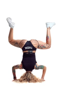 Nice blond twerk woman with tattoed body and long curly hair is posing standing on her head, isolated on white background with copy space. Attractive lady wearing in a mesh black shorts, top and white sneakers. Booty dance in studio.