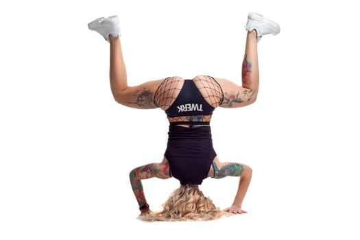 Cute blond twerk girl with tattoed body and long curly hair is posing standing on her head, isolated on white background with copy space. Sexy female wearing in a mesh black shorts, top and white sneakers. Booty dance in studio.