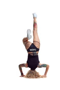 Cute blond twerk female with tattoed body and long curly hair is posing standing on her head, isolated on white background with copy space. Charming woman wearing in a mesh black shorts, top and white sneakers. Booty dance in studio.