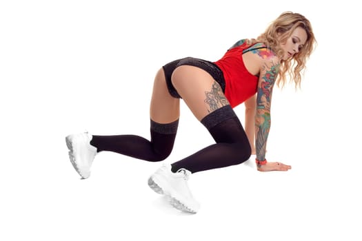 Beautiful blond lady with tattoed body and long curly hair is standing on all fours while posing isolated on white background with copy space. Young woman wearing in a black stockings and mini shorts, red top and white sneakers. Booty twerk dance in studio.