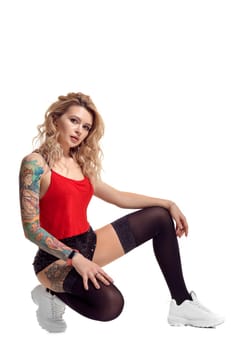 Beautiful blond woman with tattoed body and long curly hair is looking at the camera while posing isolated on white background with copy space. Young girl wearing in a black stockings and mini shorts, red top and white sneakers. Booty twerk dance in studio.