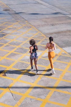 vertical rear view of two unrecognizable female friends running happy along city road in a sunny day, concept of friendship and sporty lifestyle, copy space for text