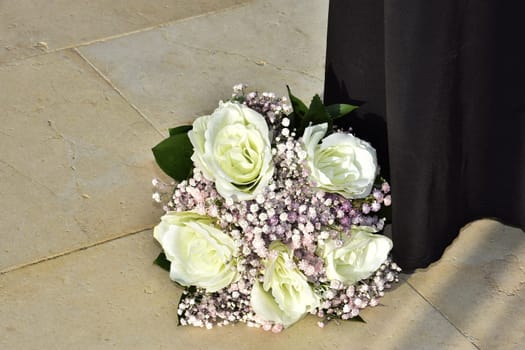 The bride's wedding bouquet of white roses and pink flowers lies on the marble yellow floor, next to the long black skirt of a standing woman. High quality photo