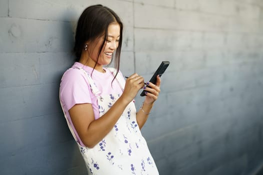 Happy Young Chinese woman using her smartphone with a pen or stylus, outdoors Asian female wearing casual clothes.