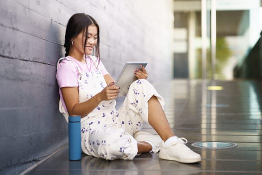 Full body glad Asian female in stylish outfit smiling and watching video on tablet while sitting on slope near thermos outside modern building in daytime