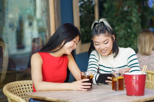 Positive young Asian female friends, in casual outfits sitting at wooden table with glasses of cold drinks and looking at smartphone screen together while watching video during meeting
