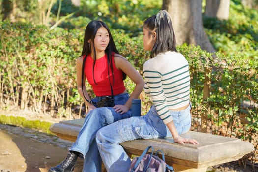 Calm young Asian female friends in casual clothes, resting on wooden bench during trip in Alhambra forest together and talking about Granada while looking at each other