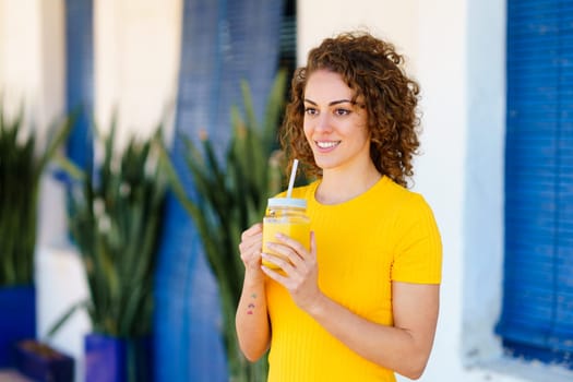 Positive young curly haired female in yellow t shirt, smiling and looking away while standing on street with glass jar of fresh healthy orange juice with straw