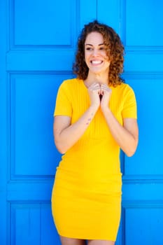 Cheerful young female in yellow dress with curly brown hair smiling and looking away against blue background on street