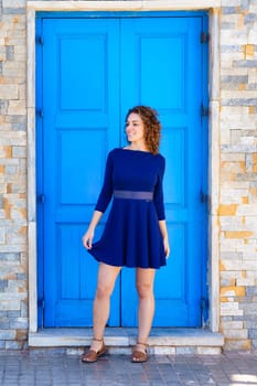 Full body of cheerful young female in blue dress smiling and looking away while standing on pavement against brick wall with bright door