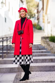 Full length of happy adult female tourist, in elegant coat and beret with photo camera on neck smiling and looking away while standing on stone stair in old town