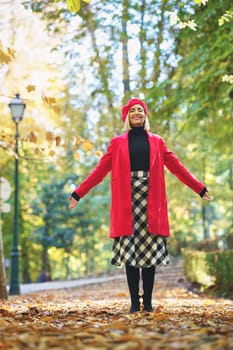 Full body of smiling female wearing elegant clothes spreading arms and throwing dry leaves on footpath in autumn park